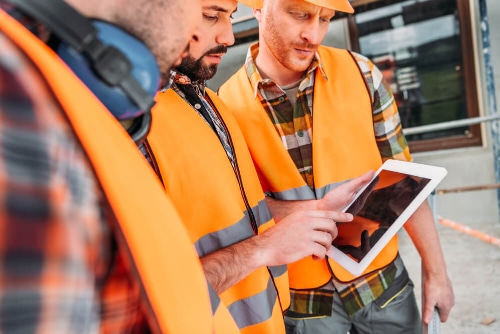 Three Smartway Builders team members in high-visibility vests and hard hats reviewing building plans on a tablet computer to ensure client expectations are met.